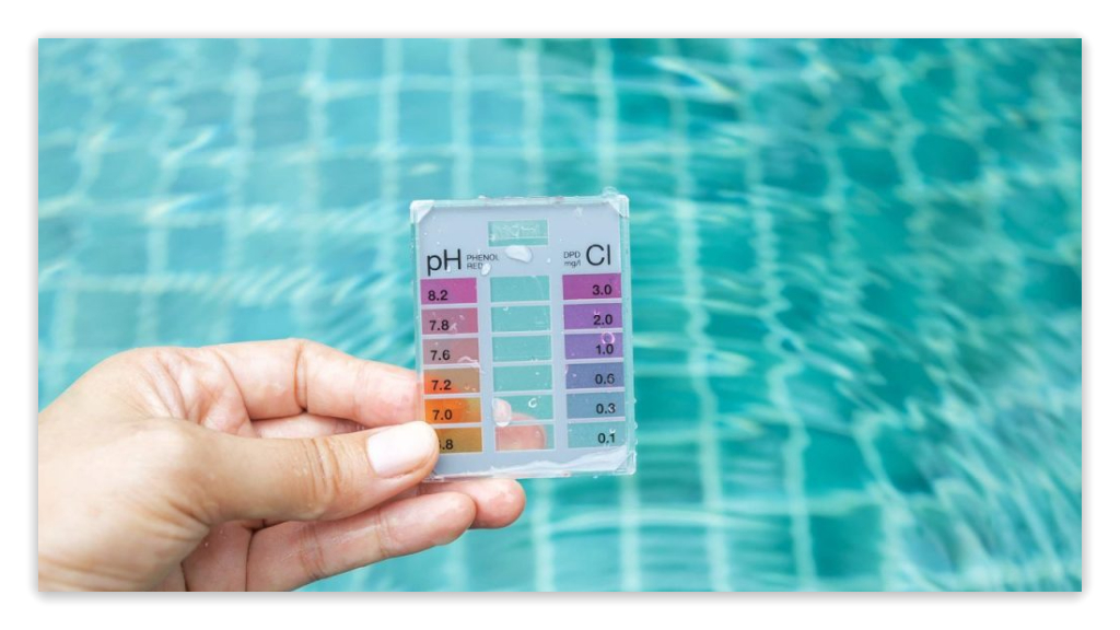 Hand holding a pH test kit above a swimming pool to check water pH levels.