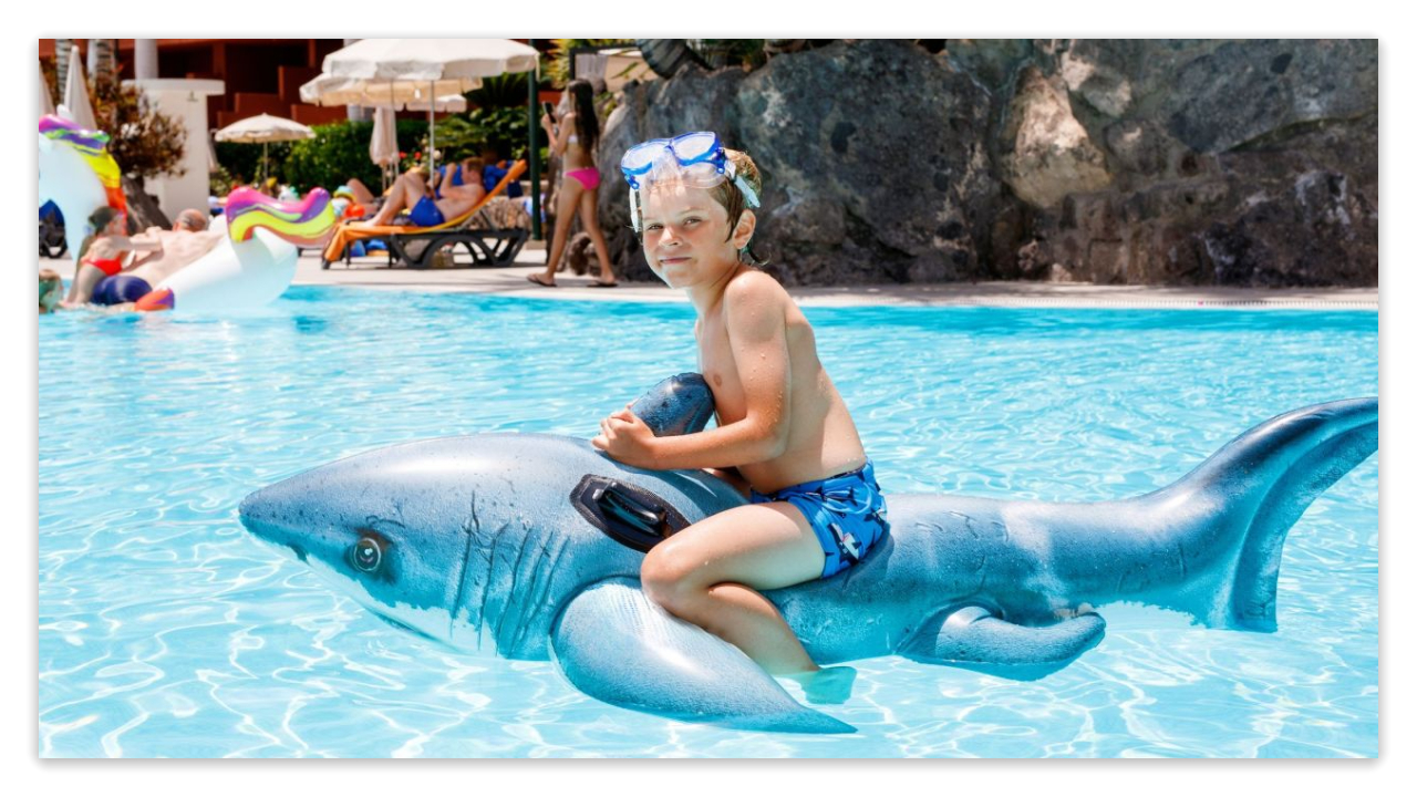 Child on an inflatable shark in a hotel pool with other guests in the background
