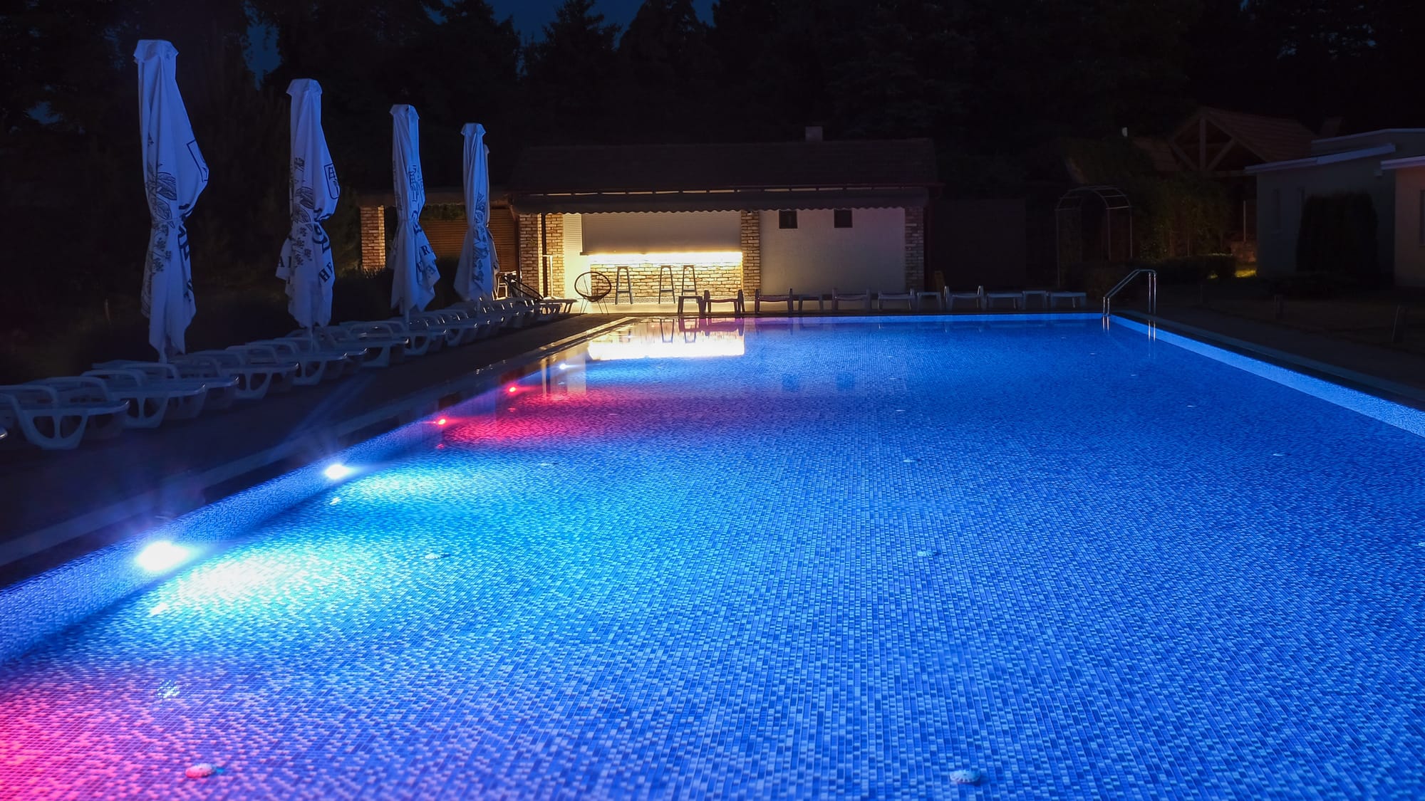 A wide shot of a swimming pool at night, its water vividly lit by striking blue and red underwater neon lights.